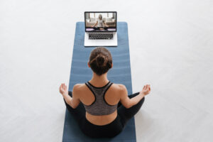 A woman sitting on top of her yoga mat in front of a laptop.